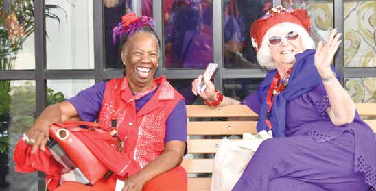 Two members in red hats and purple outfits laughing together on a bench outside a shop window