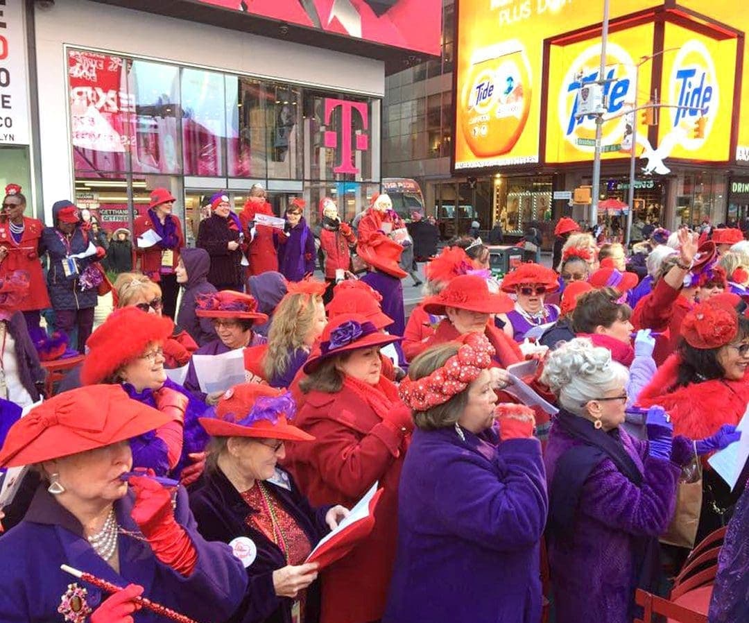 A crowd of members in red hats and purple outfits gathered outdoors at a Red Hat Society event