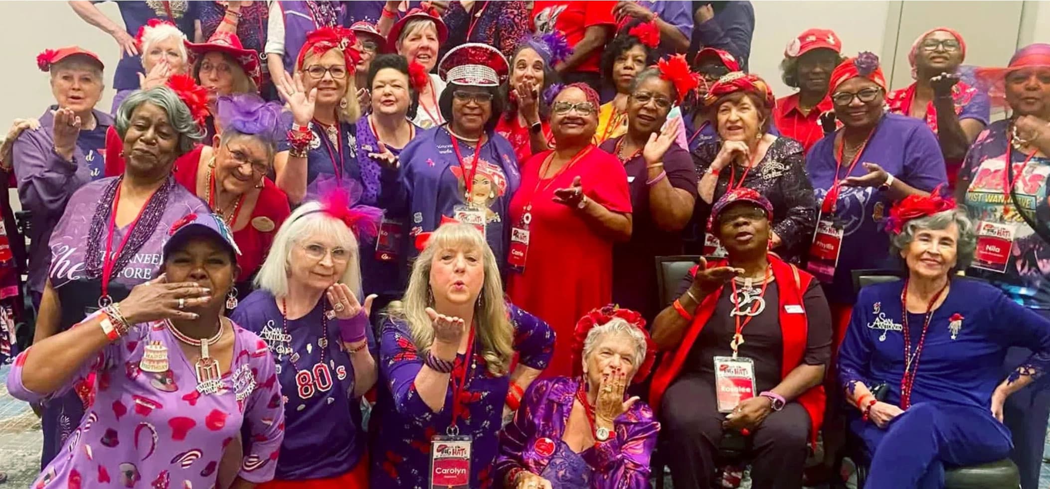 A large group of Red Hat Society members in red hats and purple outfits, gathered at a convention and blowing kisses to the camera